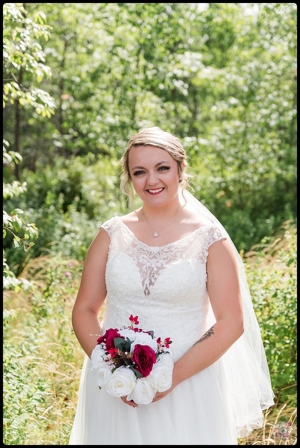 A bride in a lace wedding dress holds a red and white bouquet in a natural outdoor setting.