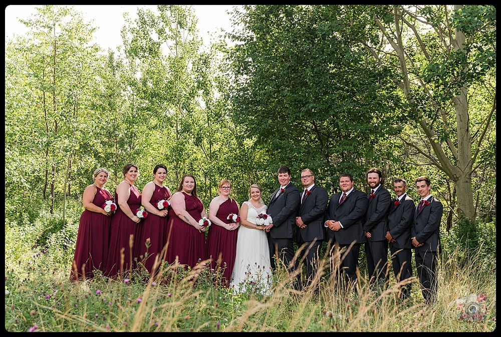 Wedding party in burgundy dresses and black suits pose together in a scenic outdoor setting.