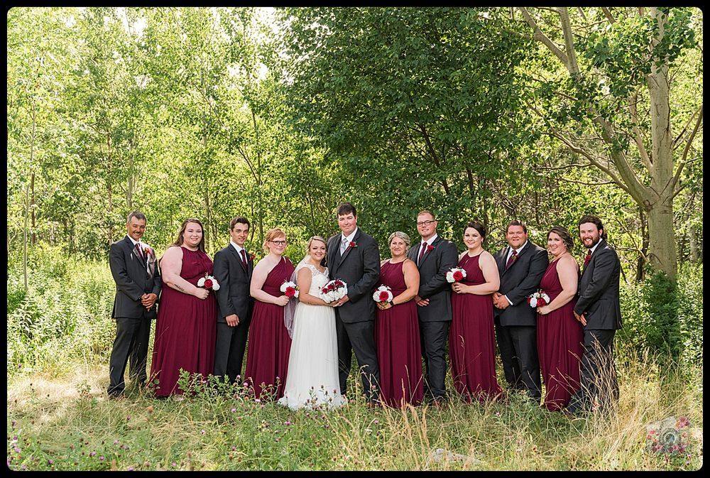 Wedding party poses in burgundy dresses and black suits against a lush green forest background.