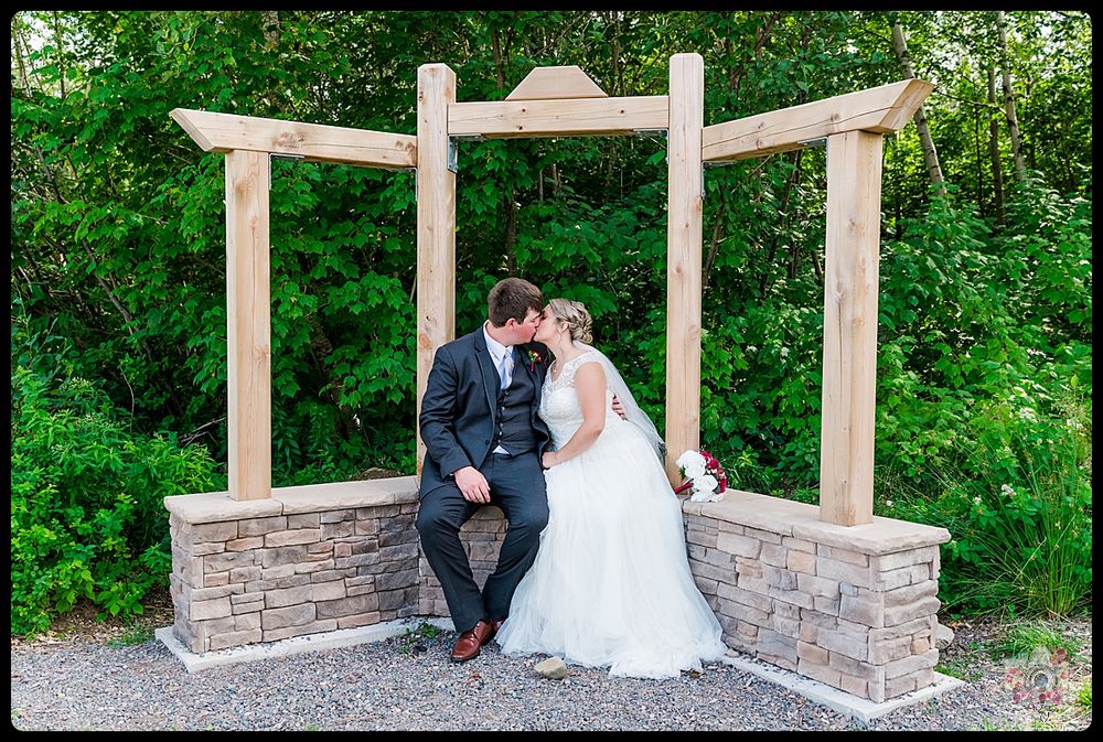 Couple sits together on stone bench under wooden pergola in garden setting.