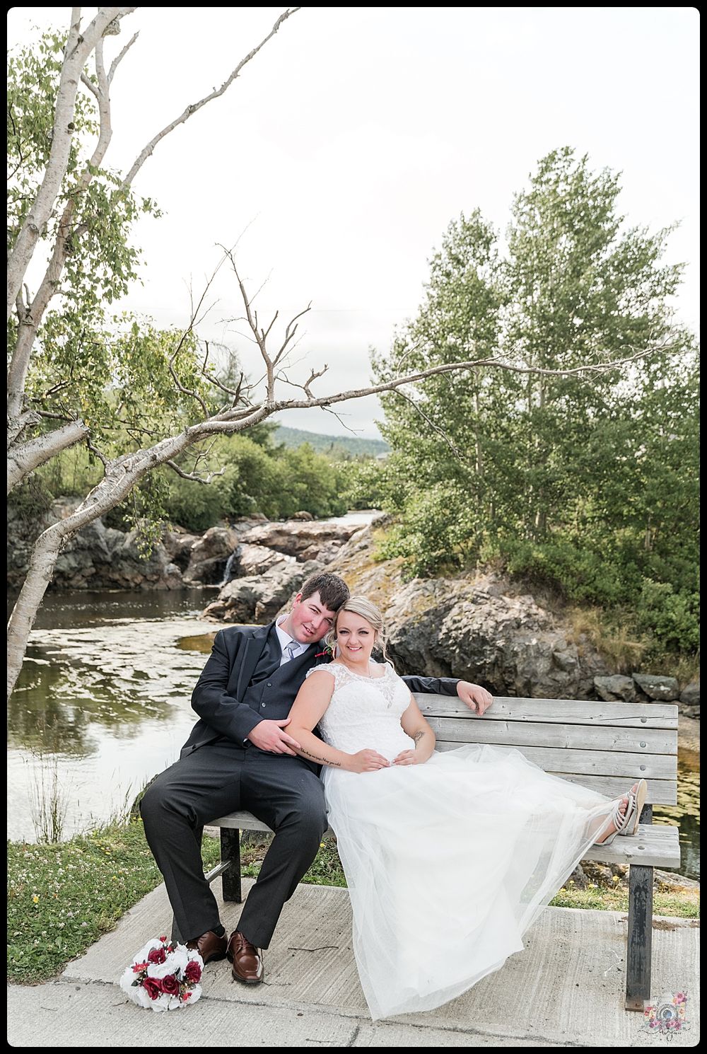 Couple relaxes on wooden bench beside stream with rocky landscape.