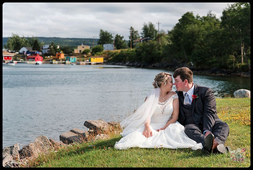 Waterfront wedding portrait with couple seated on grass near colorful harbor buildings.