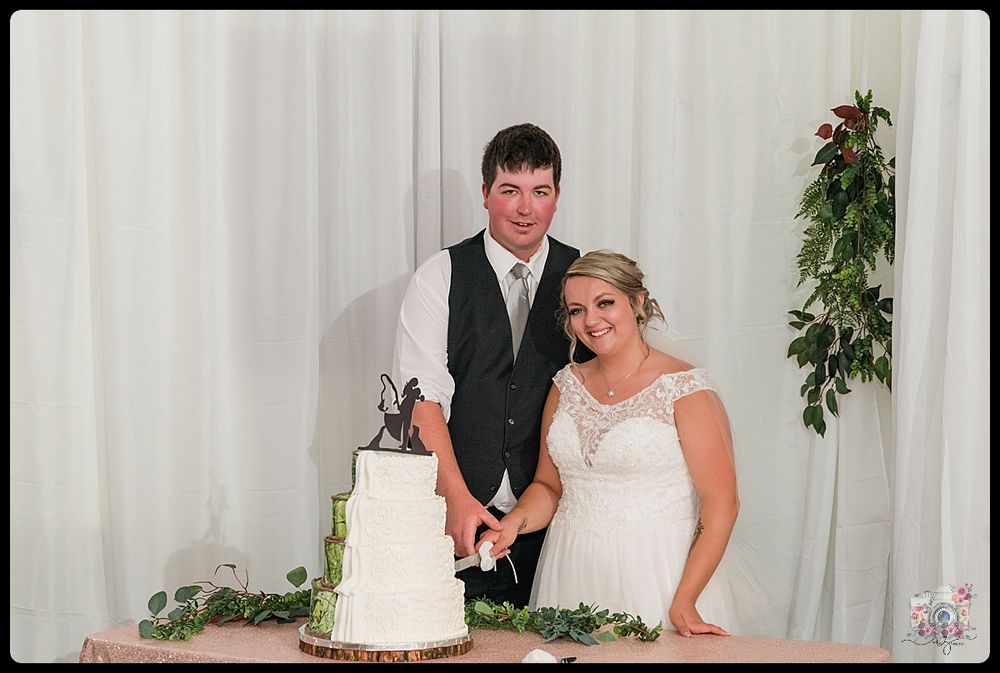 Wedding couple cutting their elegant white tiered cake at reception with greenery backdrop.