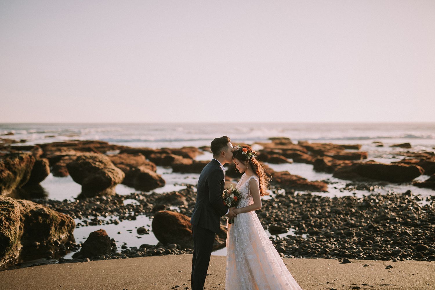Wedding couple walking along a rocky beach shoreline during sunset.