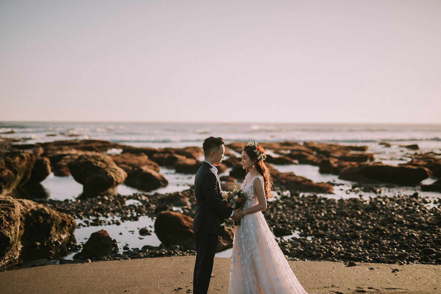 Newlyweds share intimate moments on a scenic beach with waves crashing against rocks.