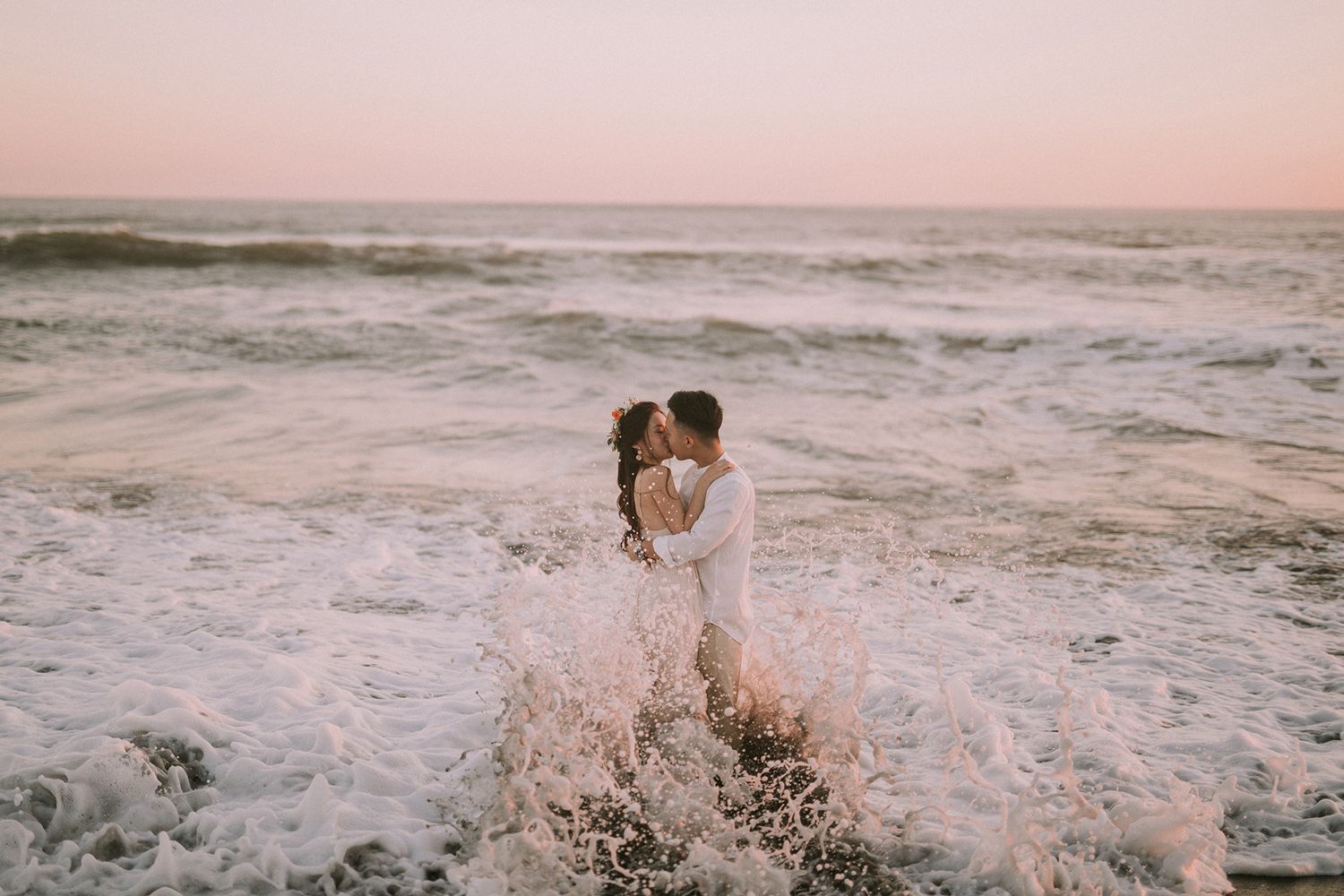 Romantic couple embraces on a misty beach at sunset with waves crashing in the background.