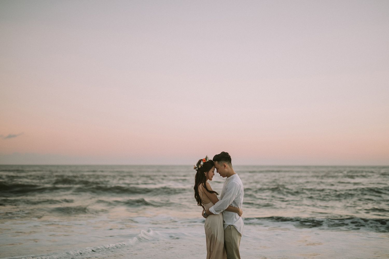 Dreamy beach photoshoot with couple silhouetted against pink sunset sky and rolling waves.