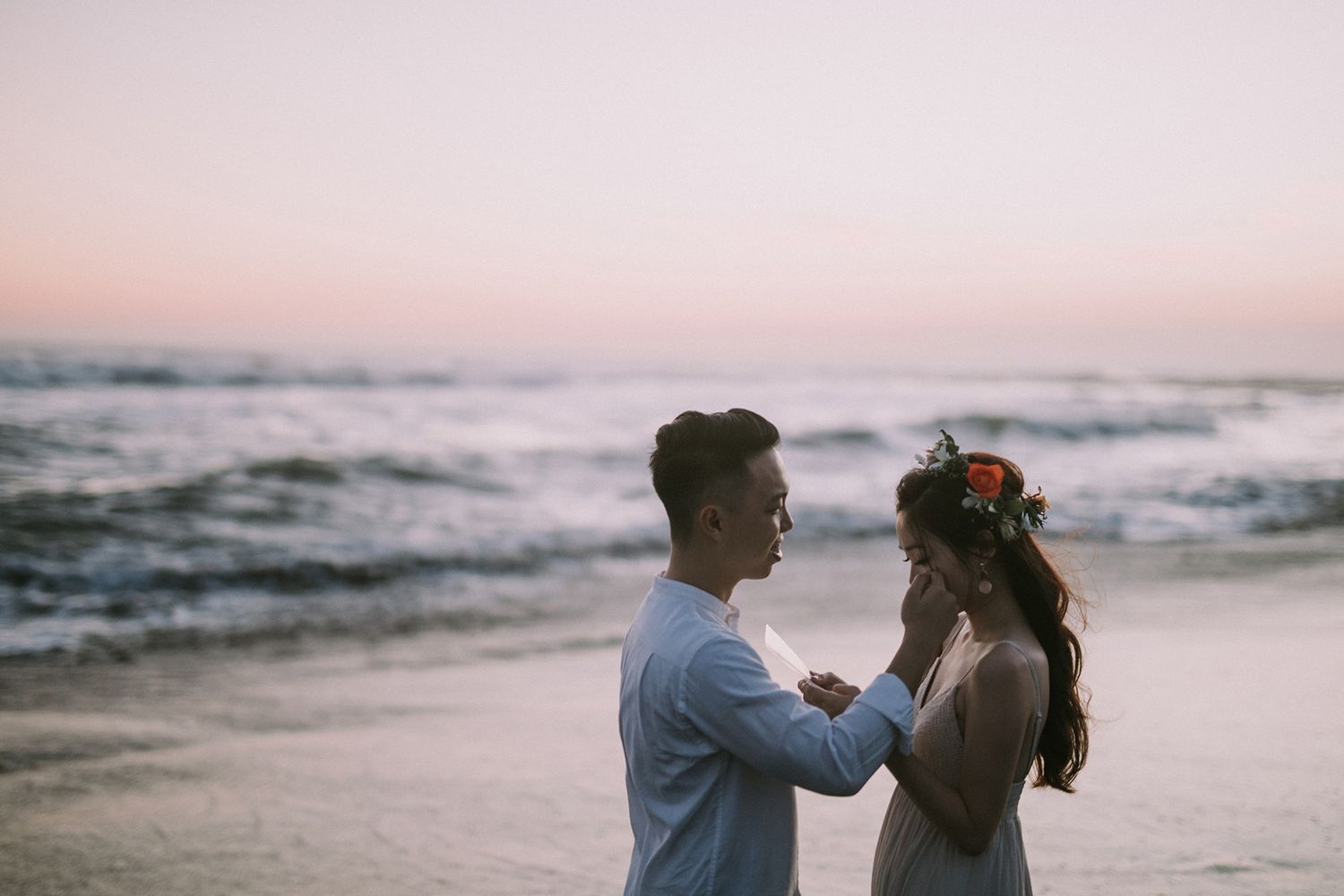 Couple in white embracing on sandy beach during sunset with waves in background.