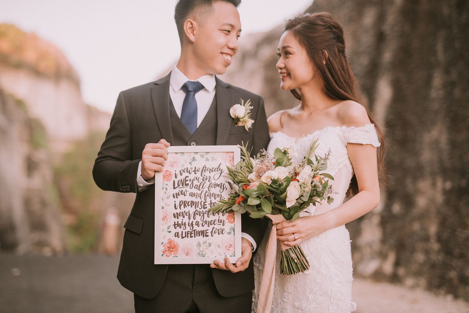 Couple poses with handwritten signs during their wedding photoshoot near coastal cliffs.