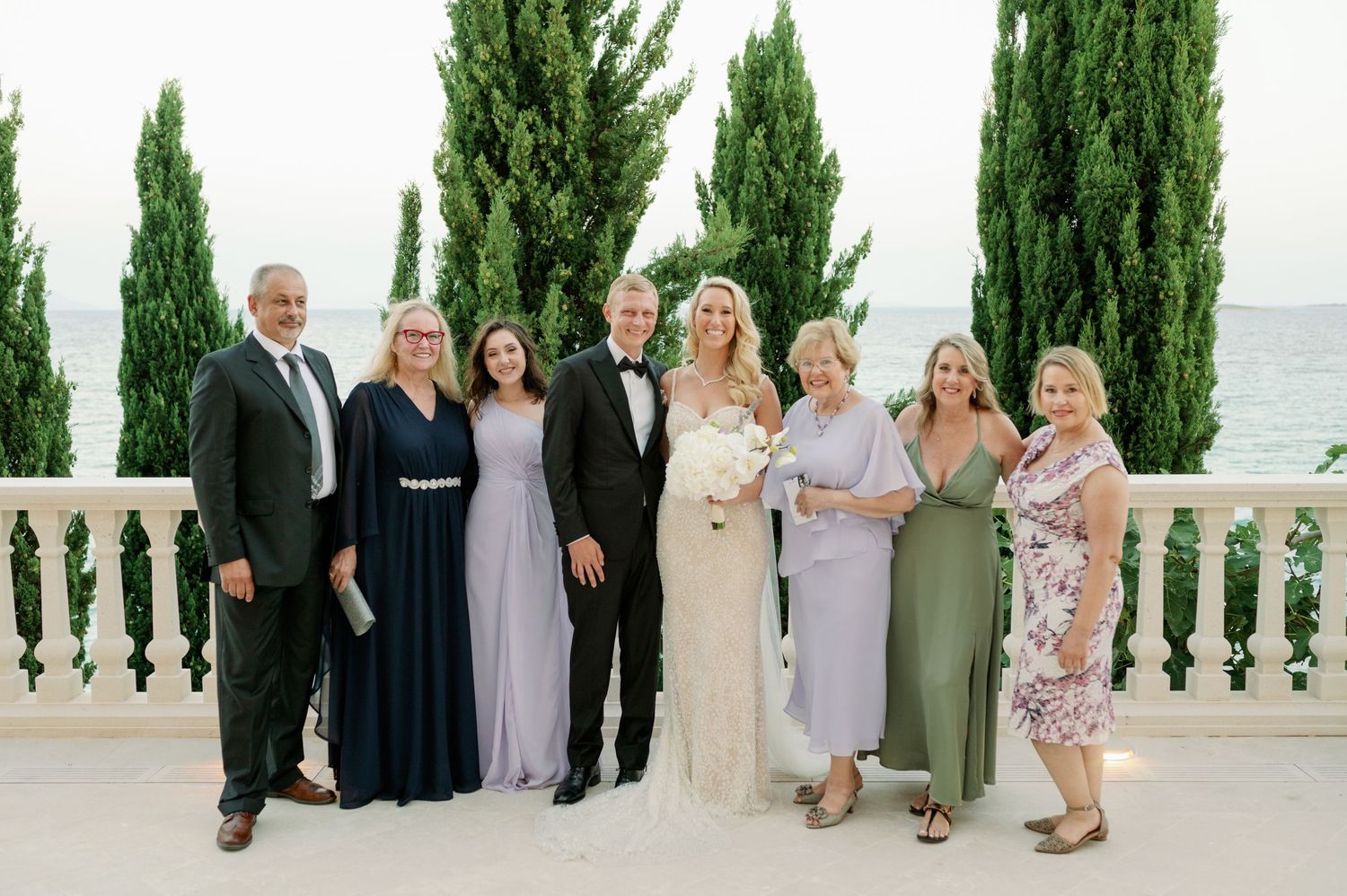 Wedding party poses for formal photo on elegant white balcony overlooking the ocean.
