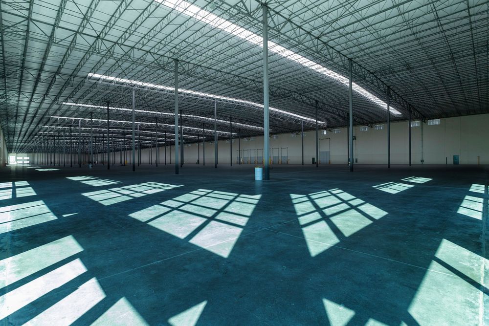 Large empty industrial warehouse interior with blue-tinted floors, steel columns, and natural light casting geometric shadows.