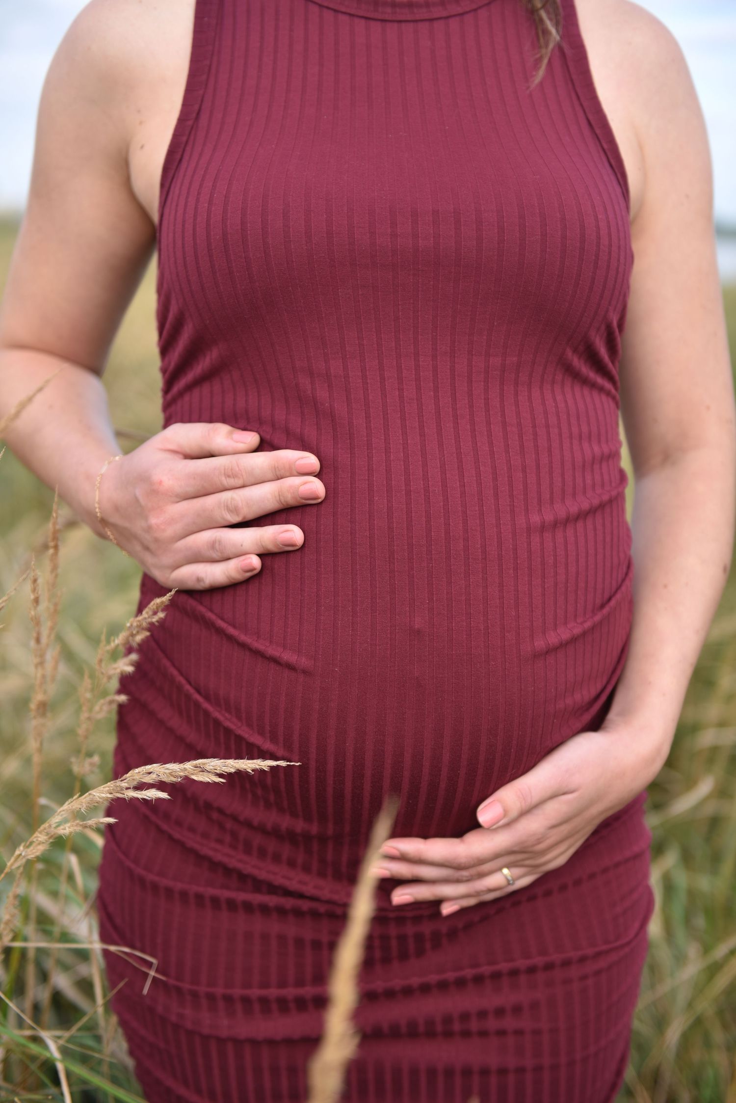 Pregnant woman in burgundy ribbed dress cradling baby bump in golden grass field.