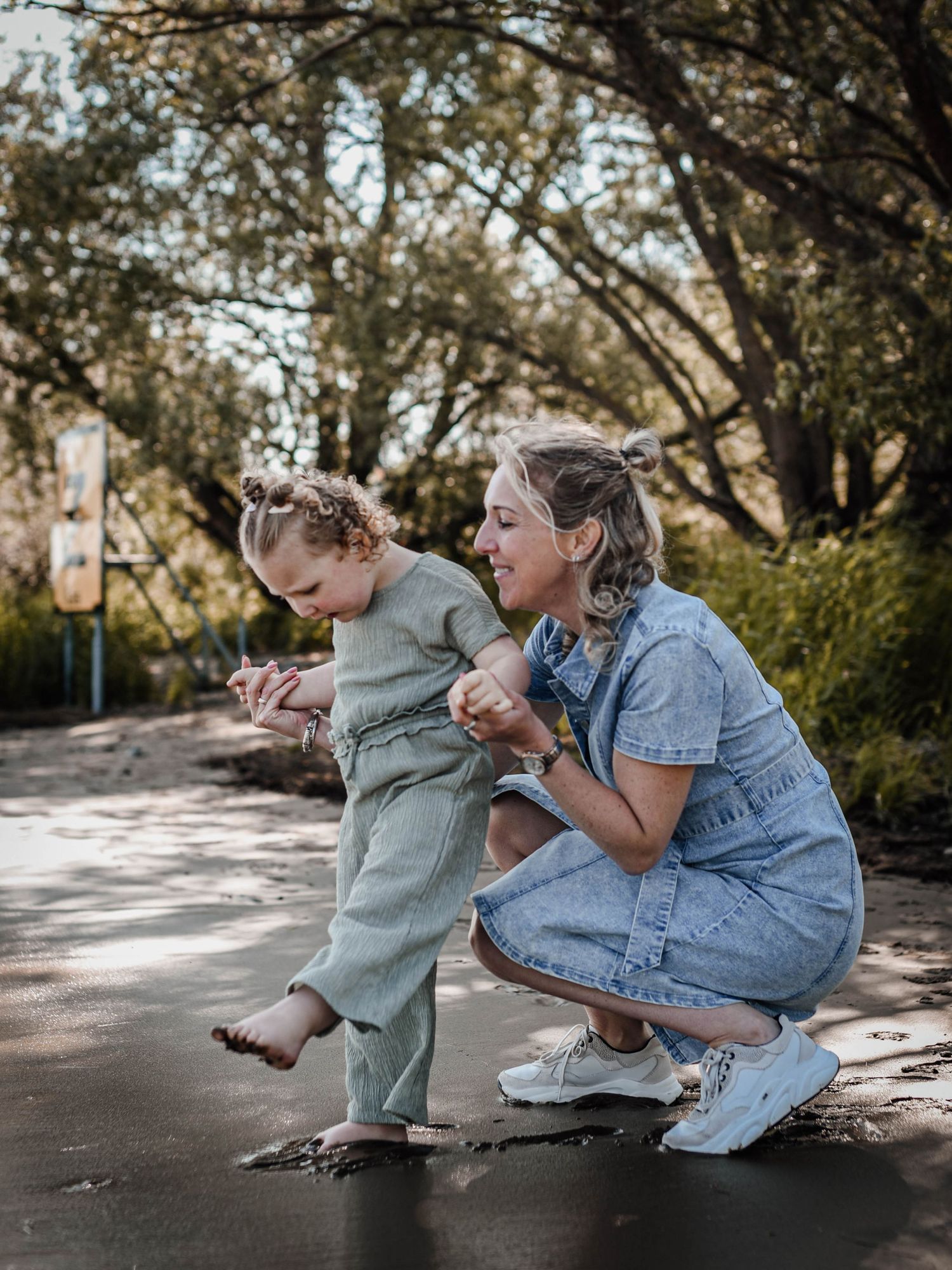 A woman in a blue dress crouches beside a toddler in a green outfit on a path surrounded by trees.