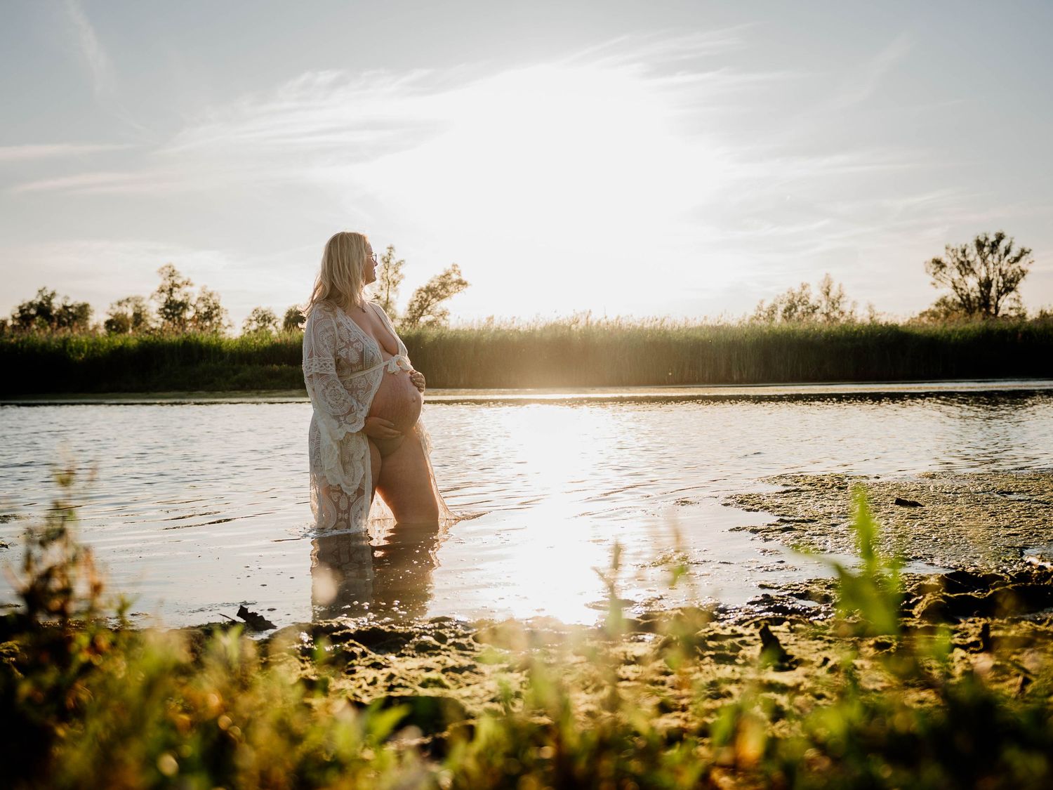 Pregnant woman standing in a sunlit river at golden hour, cradling her baby bump in a floral dress.