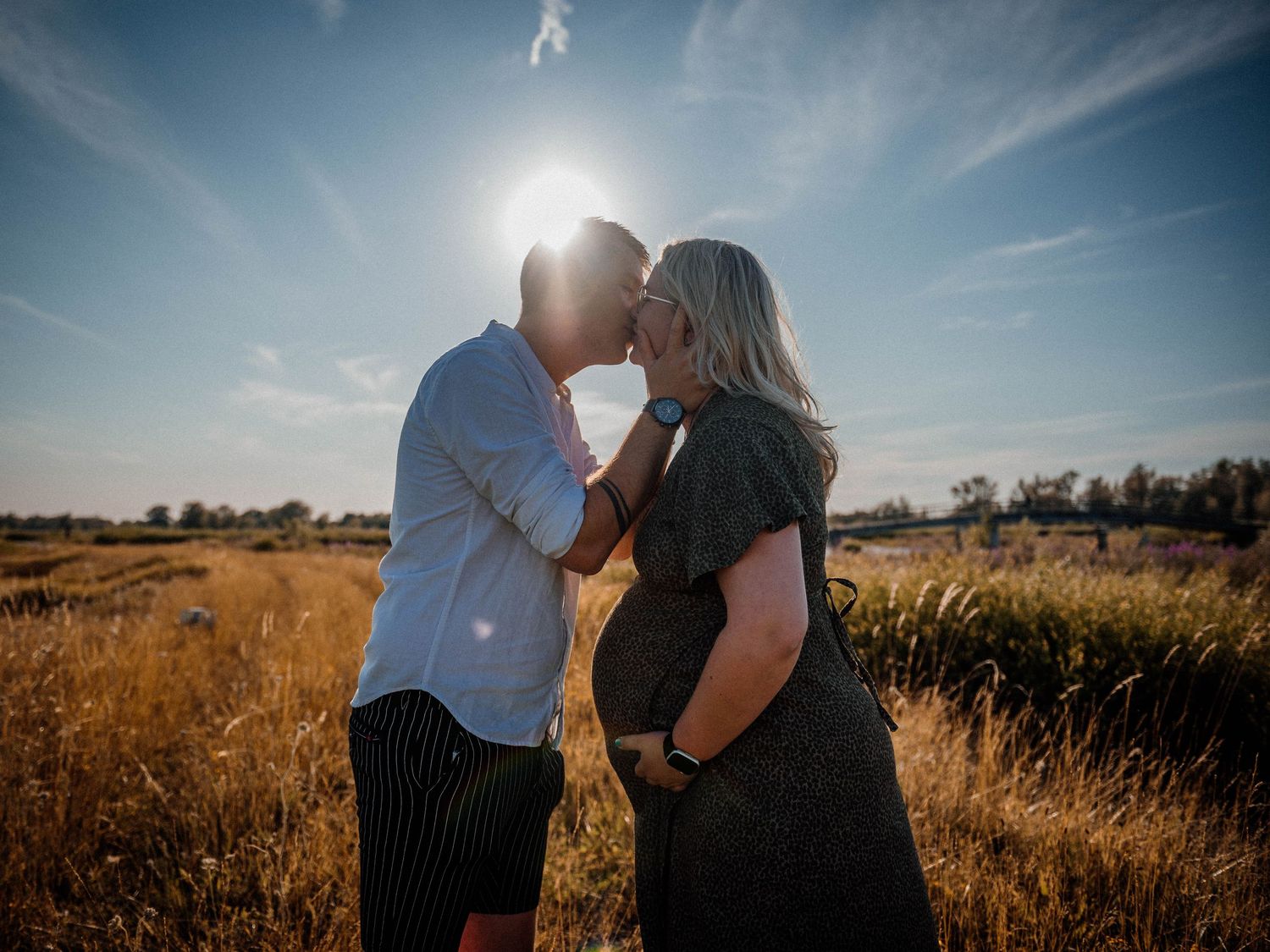 Couple kissing in golden field at sunset, pregnant woman in dark dress embraced by partner in white shirt.