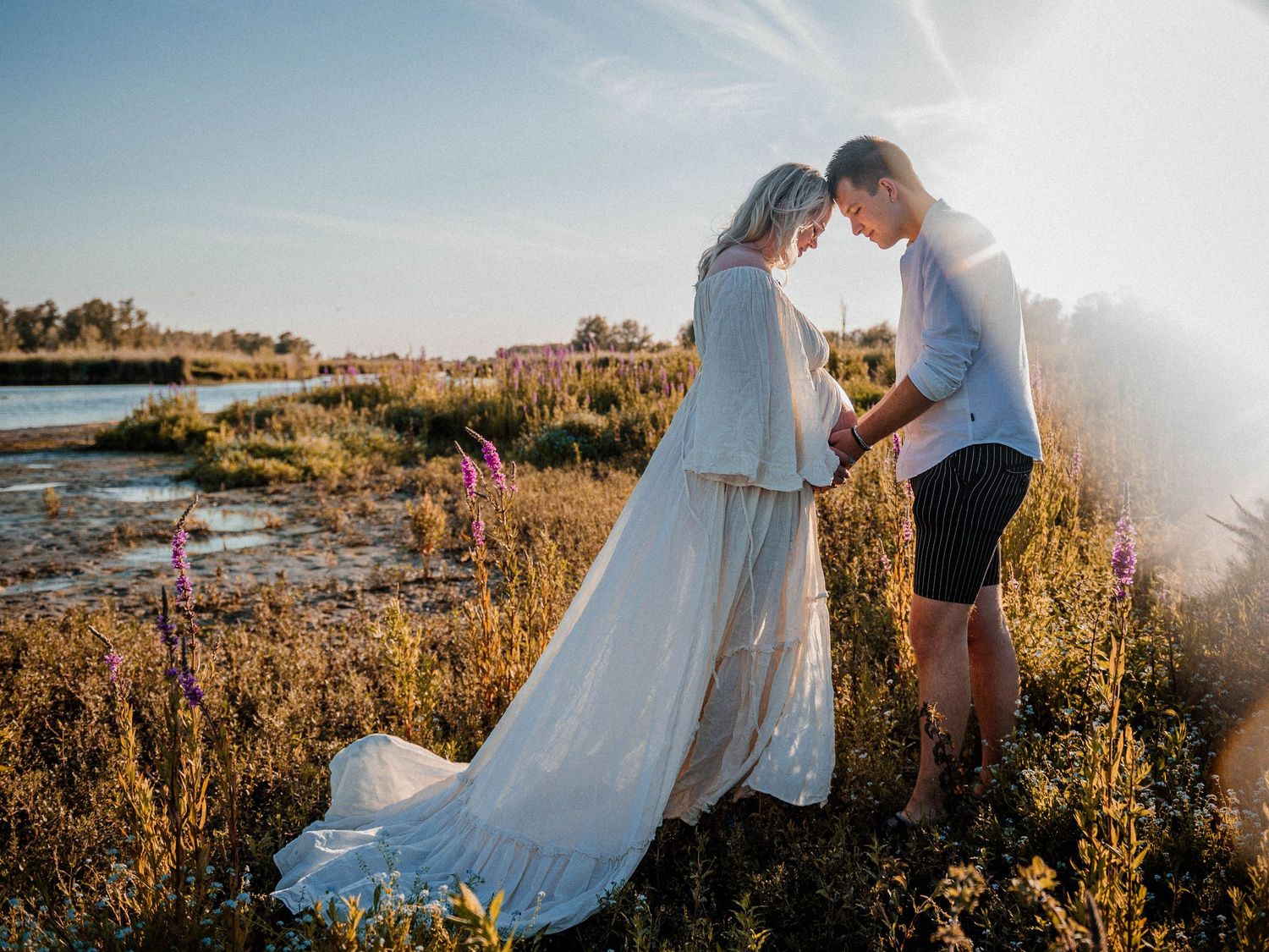 Couple in wedding attire sharing a romantic moment in a sunlit wildflower field near a river.