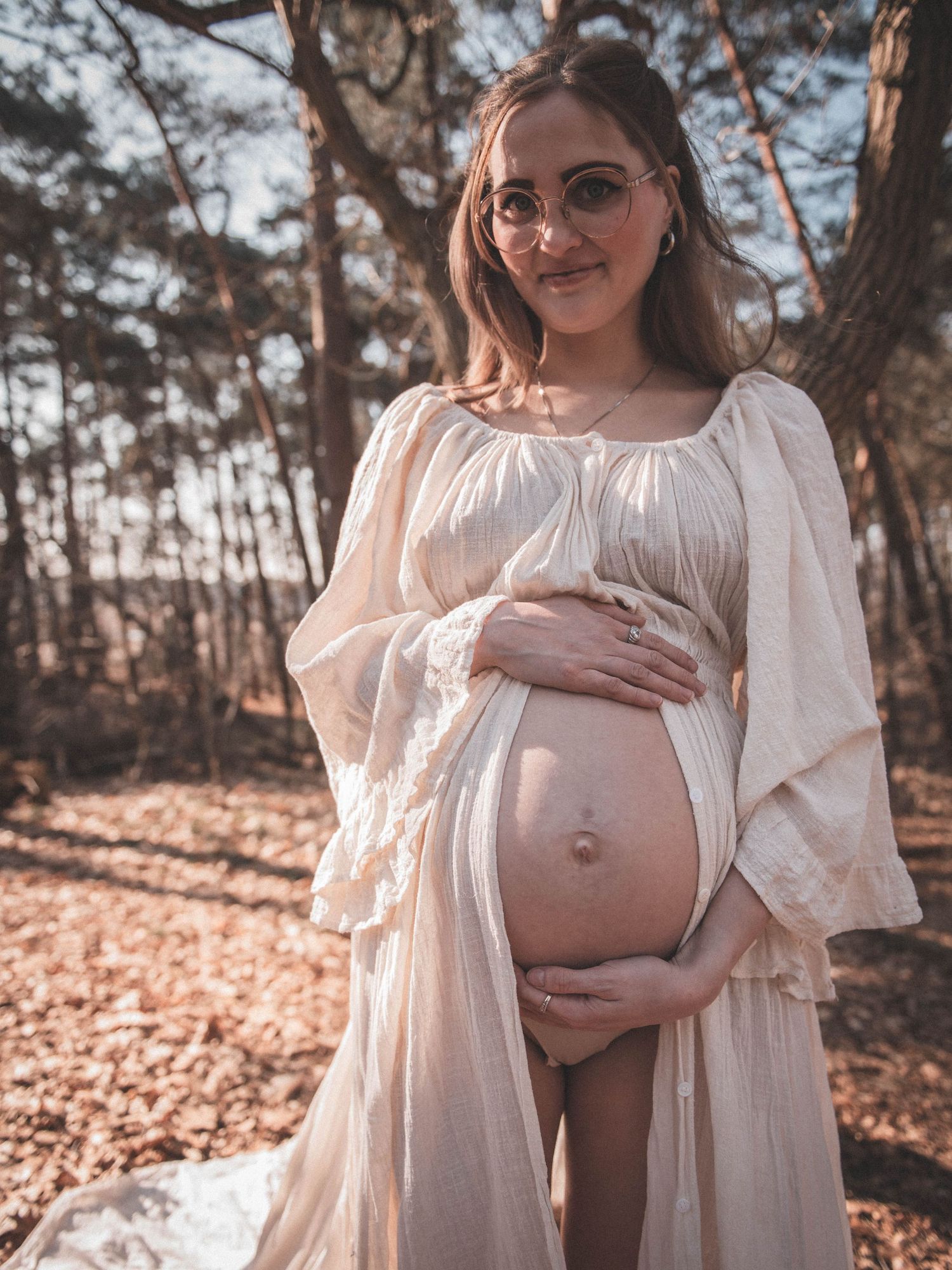 Pregnant woman in white boho dress cradling baby bump outdoors in sunlit forest during maternity photoshoot.