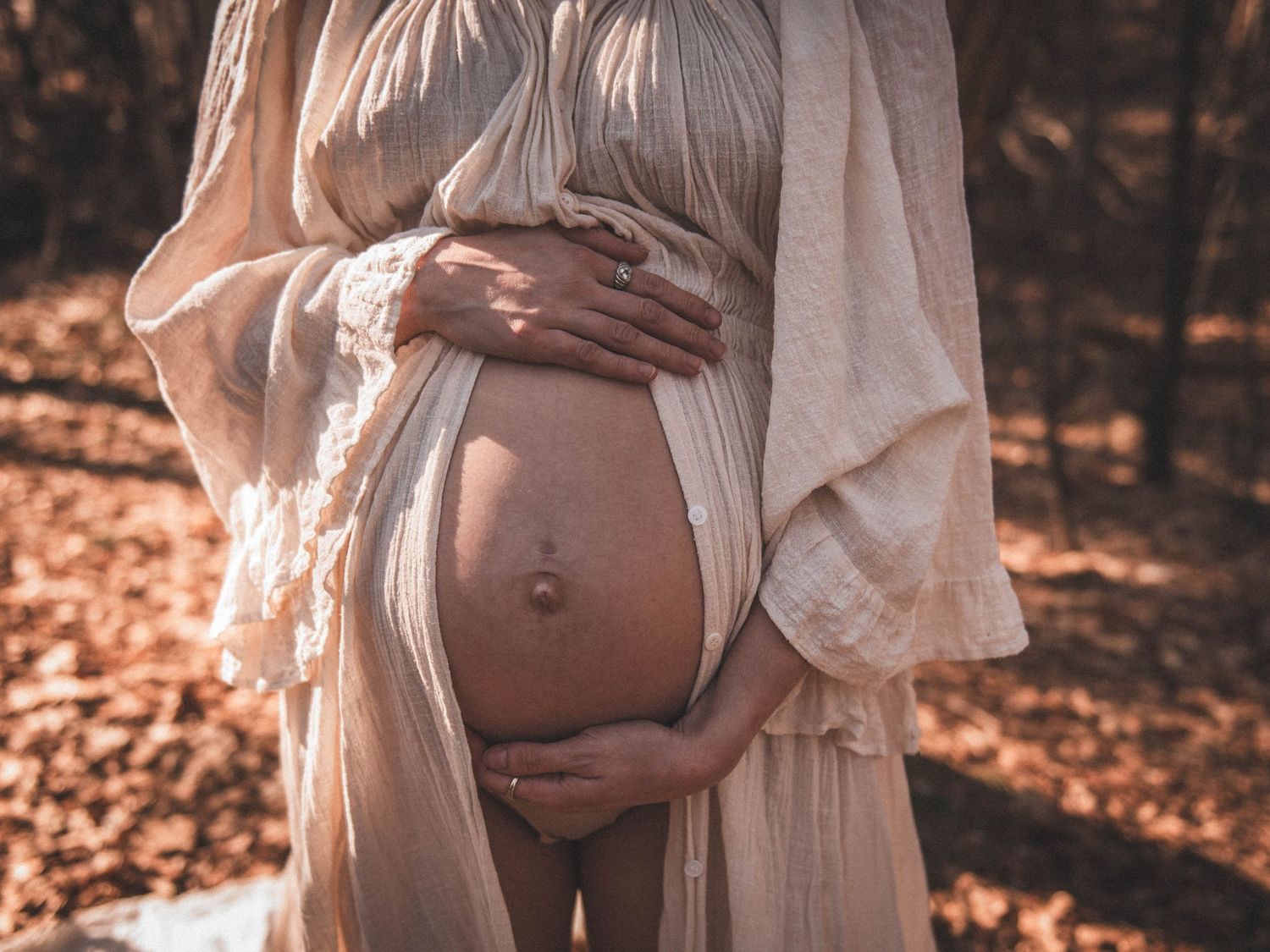 Pregnant woman in flowing beige lace robe cradling her bare baby bump outdoors in autumn forest setting.