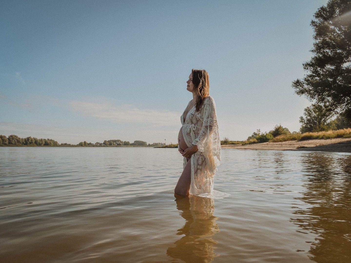 Pregnant woman in white lace dress standing in shallow river water under sunny sky with trees in background.