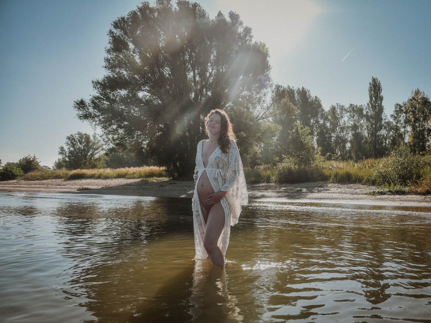 Woman in white lace cover-up wading in shallow river with sunlight streaming through trees on sunny day.