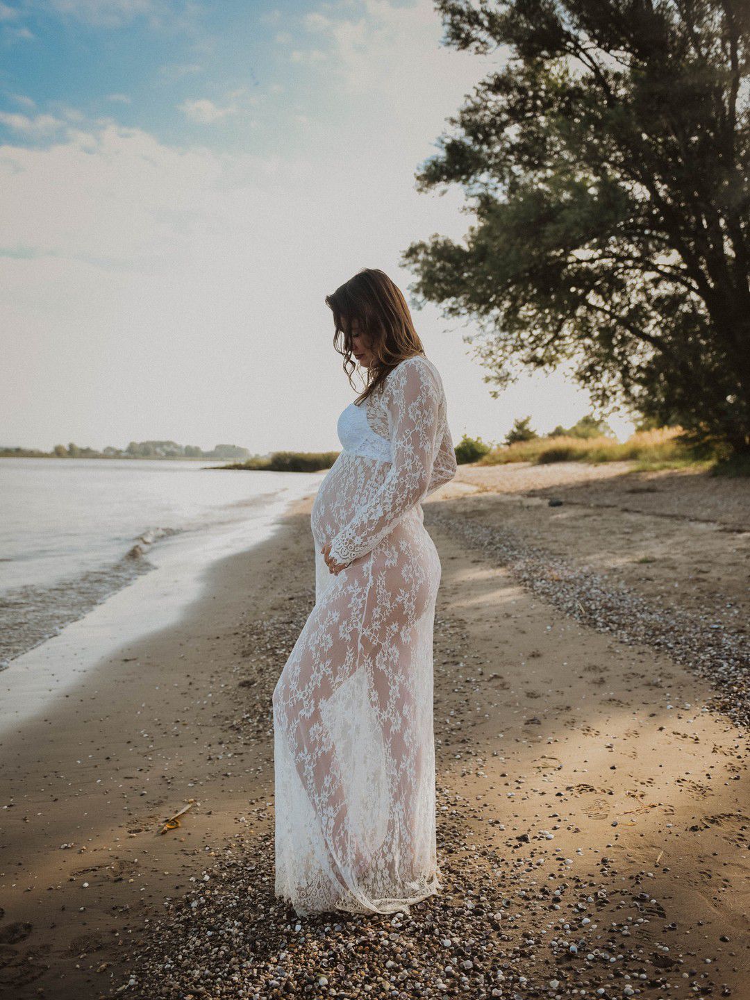Pregnant woman in white lace gown cradling baby bump on sandy beach at golden hour.