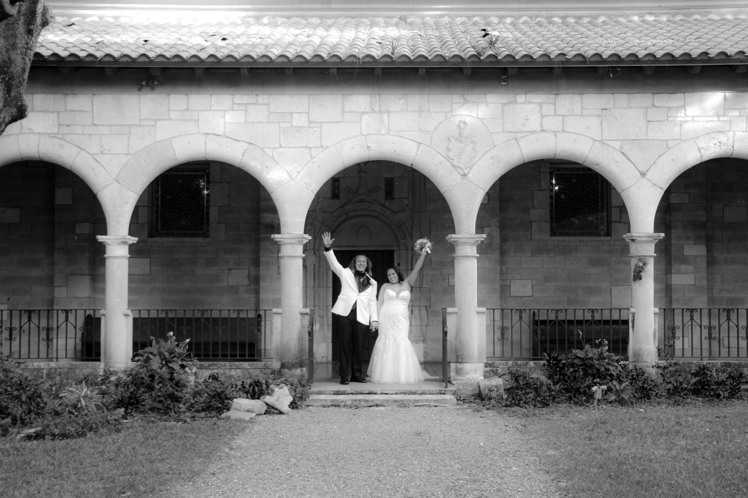 Black and white photo of newlyweds celebrating under stone arches at historic venue.