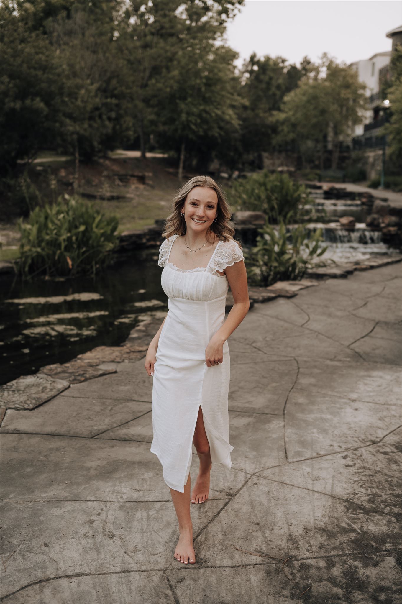 A person in a white dress walks barefoot along a stone path by a landscaped pond.