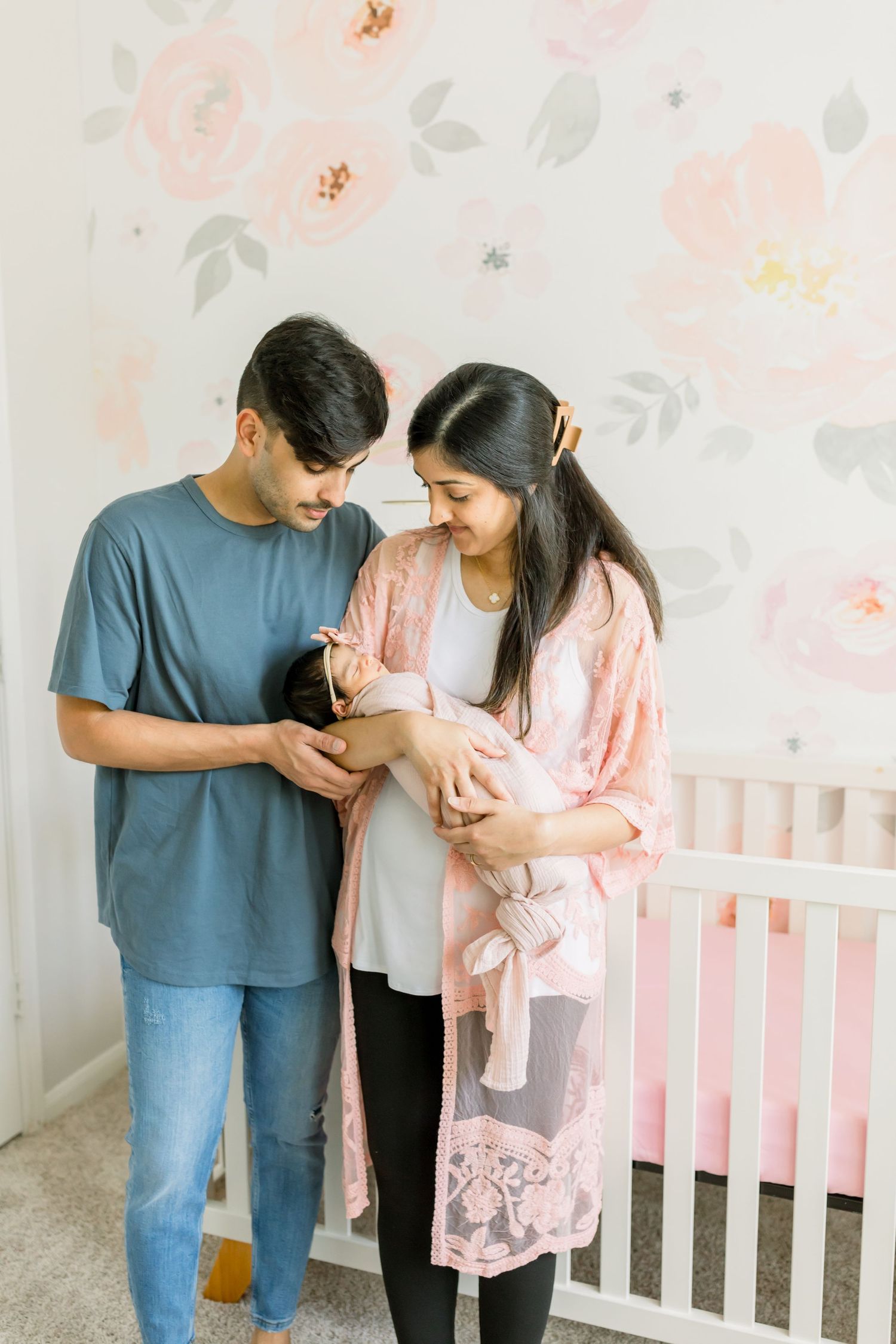 Parents standing by pink crib admiring their swaddled newborn in nursery setting.