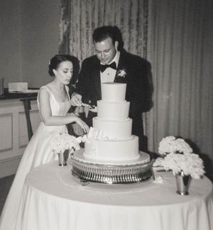 Vintage black and white photo of bride and groom cutting tiered white wedding cake at reception.
