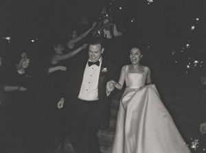 Joyful bride and groom in black and white photo walking through sparkler send-off at night.