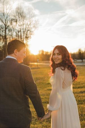 Smiling red-haired bride in white dress holding groom's hand walking through a sunlit field at sunset.