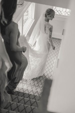 A black and white photo of a bride in a lace gown and veil descending a staircase with bridesmaids.