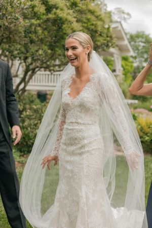 Radiant bride in long-sleeve lace wedding gown and cathedral veil laughing joyfully outdoors.