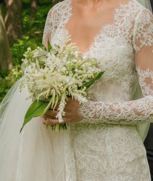 Close-up of bride holding elegant white lily of the valley bouquet in lace long-sleeve gown.