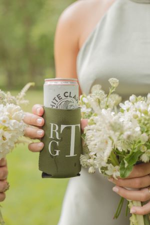 A bridesmaid holds a monogrammed green koozie with a White Claw and a white floral bouquet outdoors.