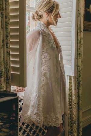 Bride in lace wedding robe standing by window with shutters, smiling softly in elegant bridal preparation moment.