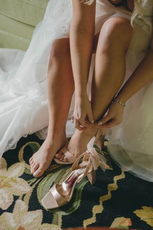 Bride putting on blush satin heels with bow detail, assisted by bridesmaid on floral carpet before wedding.