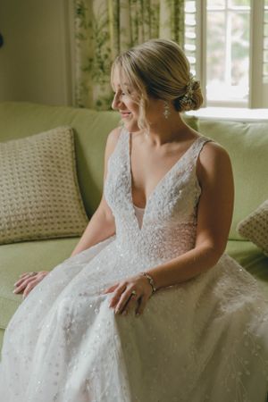 Smiling bride in sparkly deep V-neck wedding gown sitting on green sofa with elegant updo and earrings.