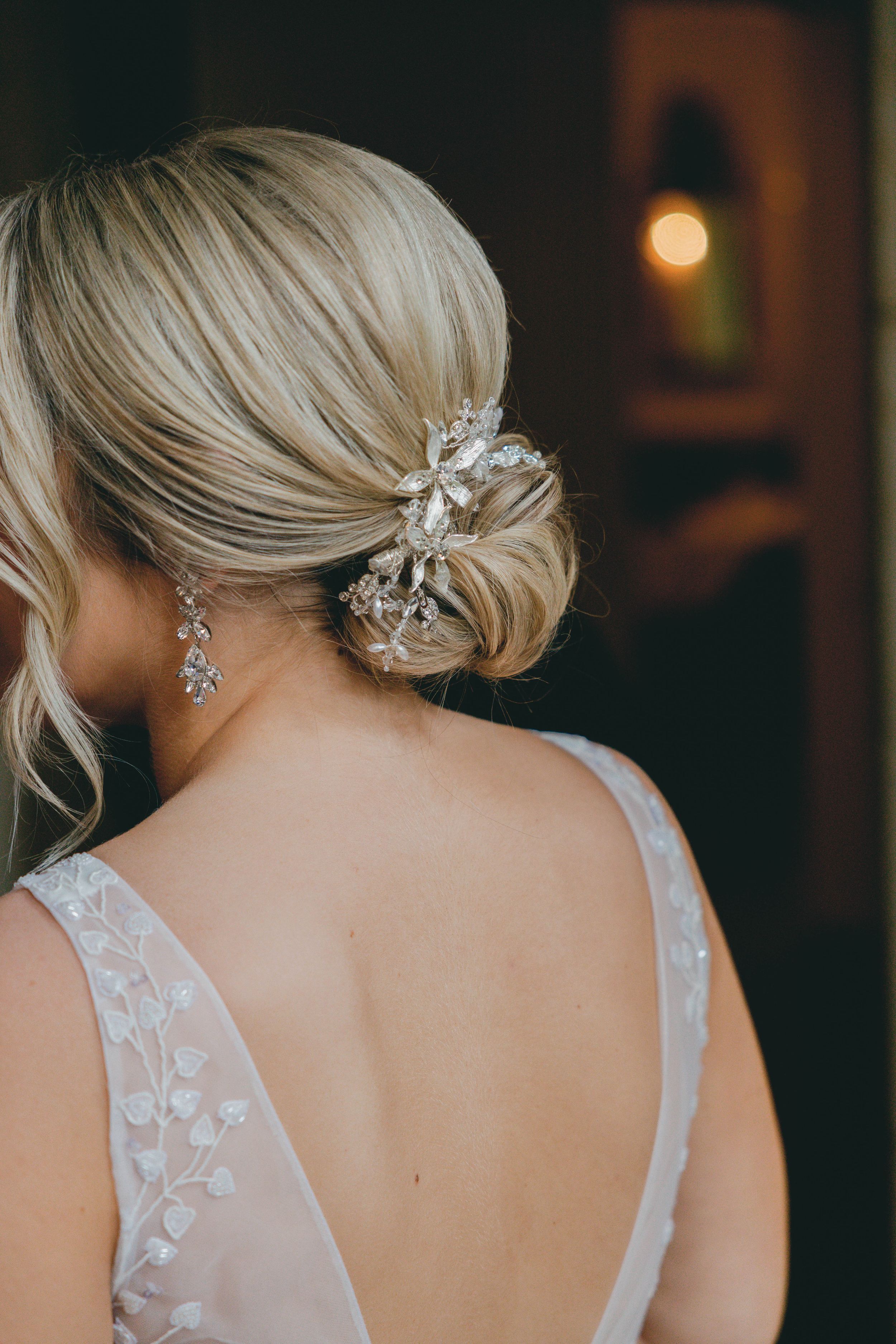 Elegant bridal updo with crystal hair accessory and drop earrings on blonde bride in backless lace wedding gown.