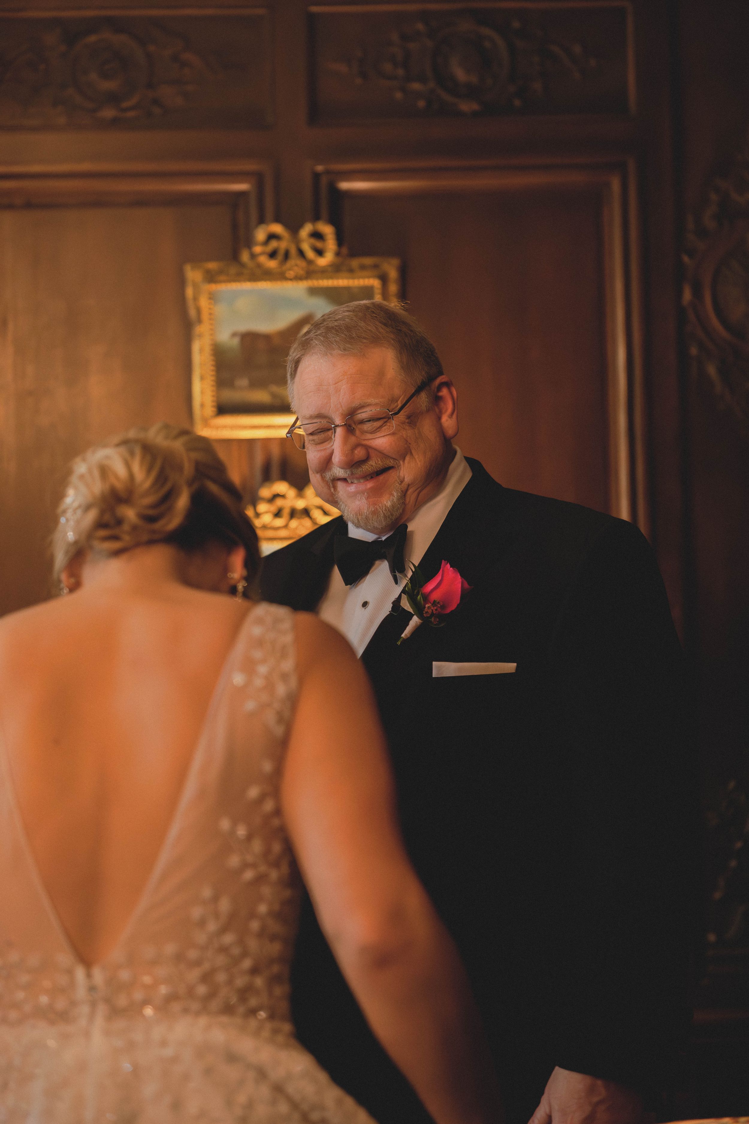 Emotional father smiling at bride during first look in ornate wood-paneled room with rose boutonniere.