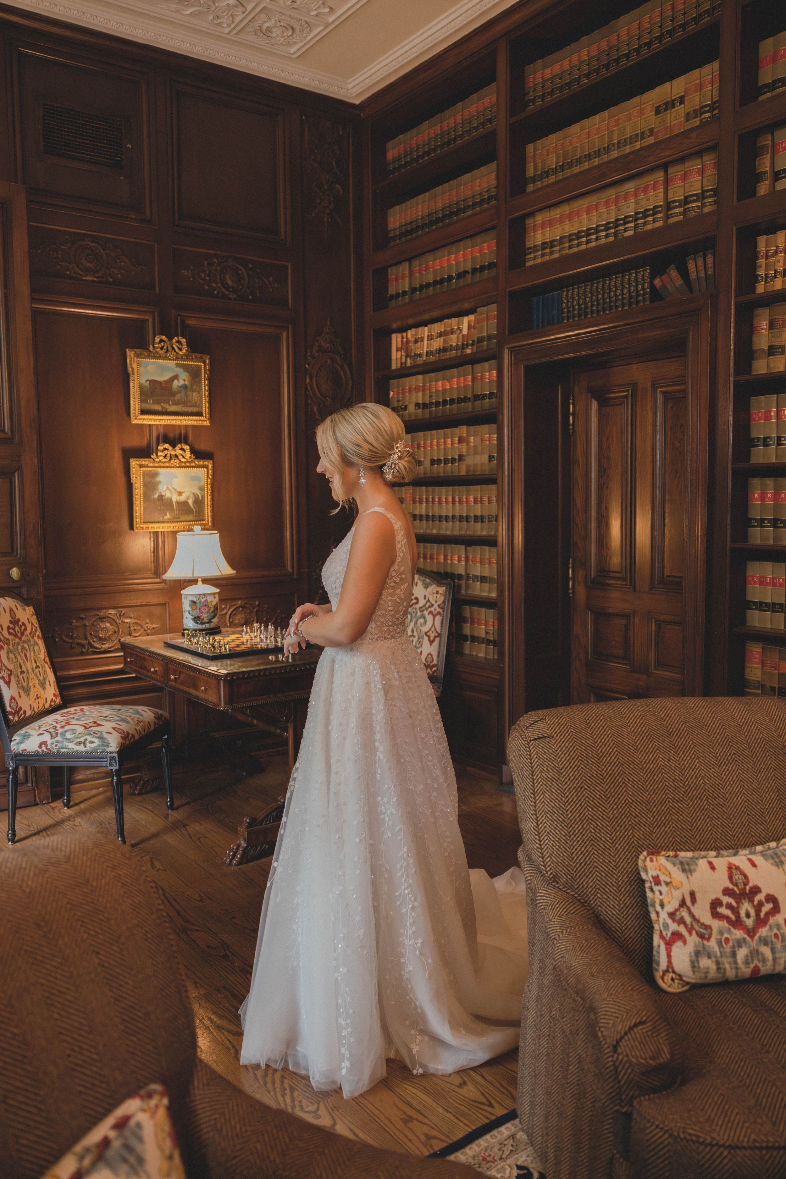 Blonde bride in sparkly A-line wedding dress standing in wood-paneled library with bookshelves and warm lighting.