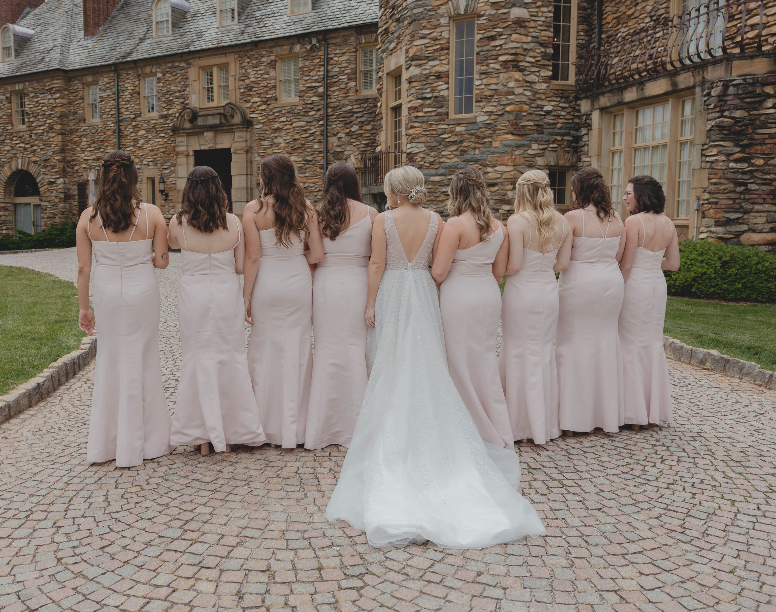 Bride and bridesmaids in blush pink gowns viewed from behind on cobblestone courtyard of stone castle venue.