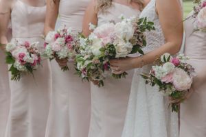 Bridesmaids in blush pink gowns holding lush pink and white peony bouquets with greenery at outdoor wedding.