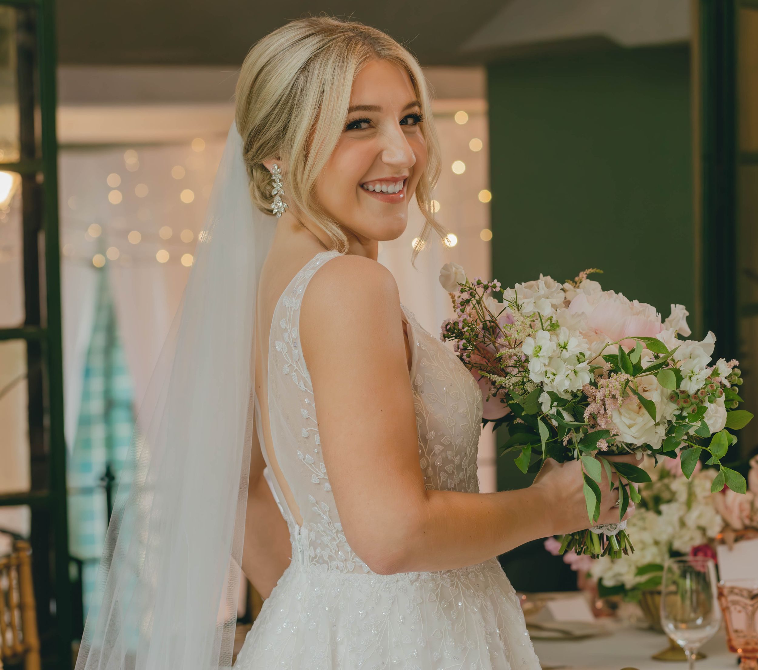 Smiling blonde bride in lace wedding dress holding pink and white floral bouquet with veil and chandelier earrings.