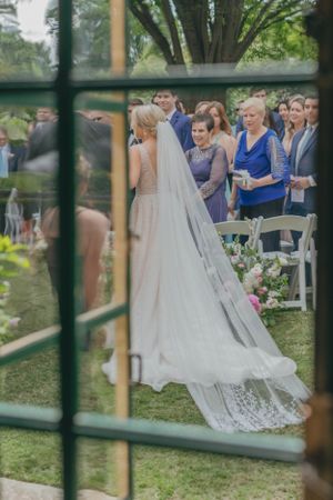 Bride in lace wedding gown with cathedral veil viewed through glass door at outdoor garden ceremony.