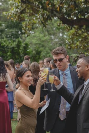 Wedding guests toasting with champagne and mimosas outdoors under trees during cocktail hour celebration.