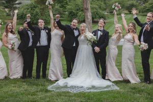 Joyful wedding party in blush and black formal wear celebrating on green lawn with arms raised in cheer.