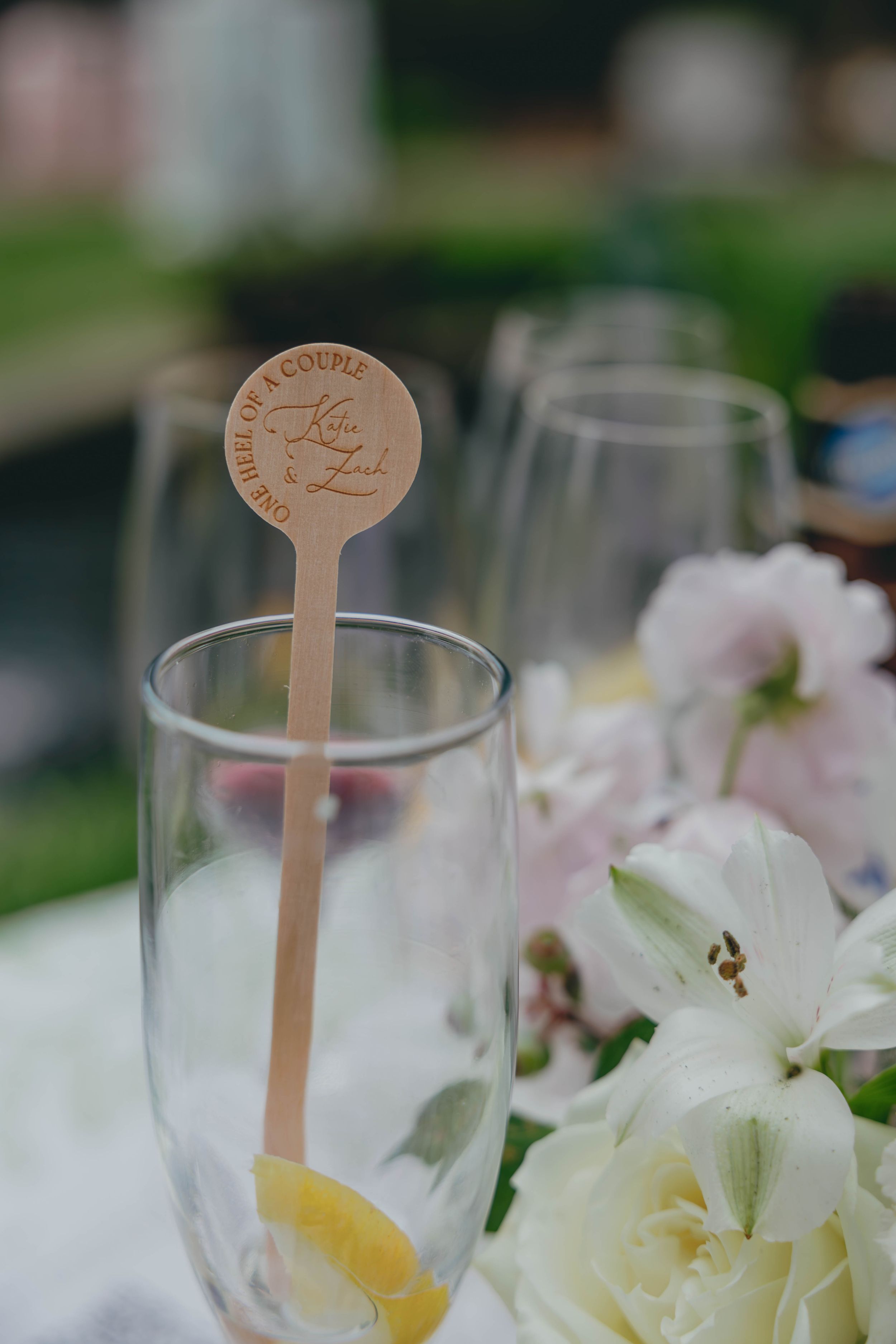 Custom engraved wooden cocktail stirrer in champagne flute with white flowers at elegant outdoor wedding reception.
