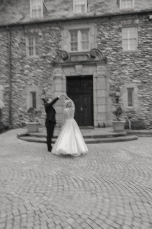 Black and white photo of bride and groom dancing and spinning on cobblestone courtyard outside stone manor.