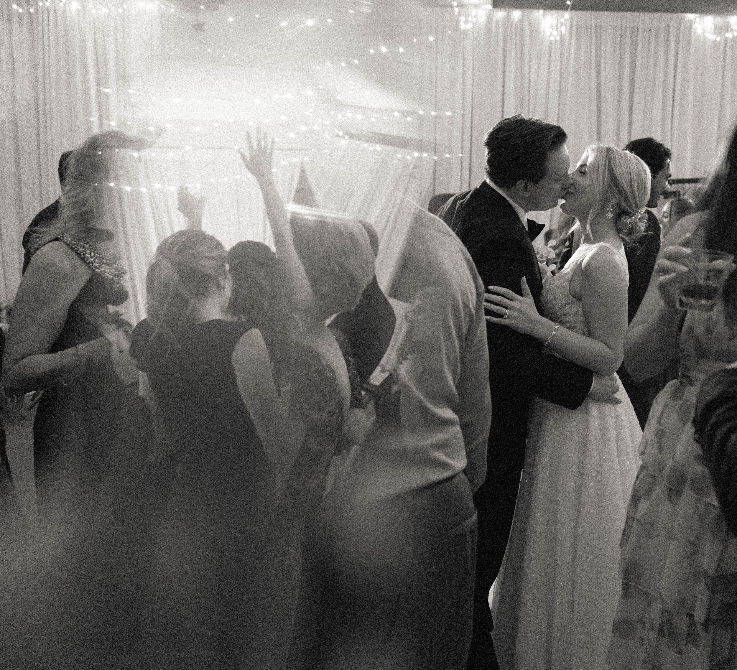 Black and white romantic photo of bride and groom kissing on crowded wedding reception dance floor.